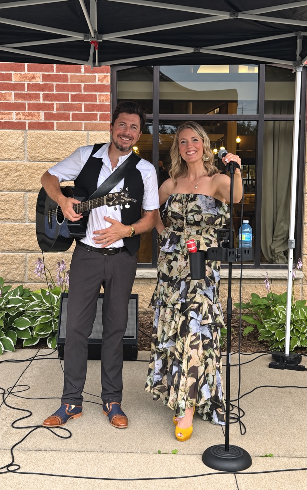 Alex with his acoustic guitar and Mia at the microphone, smiling together at an outdoor performance